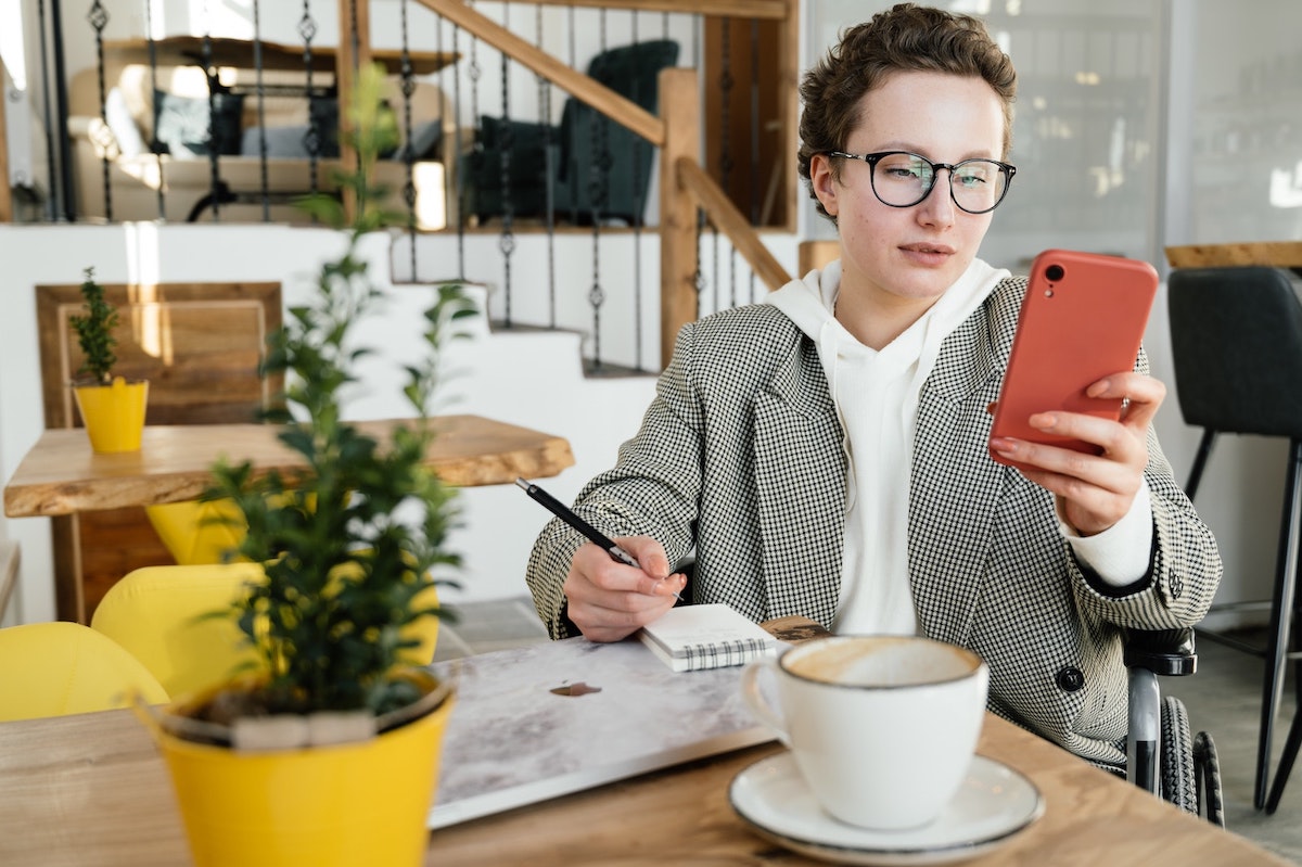 A person at a table looking at their phone and taking notes on a notepad while drinking coffee